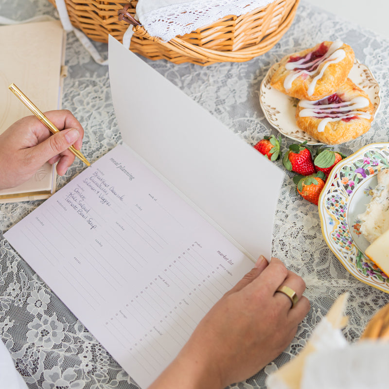 Person writing in a weekly meal planner with pastries and strawberries on a lace tablecloth.