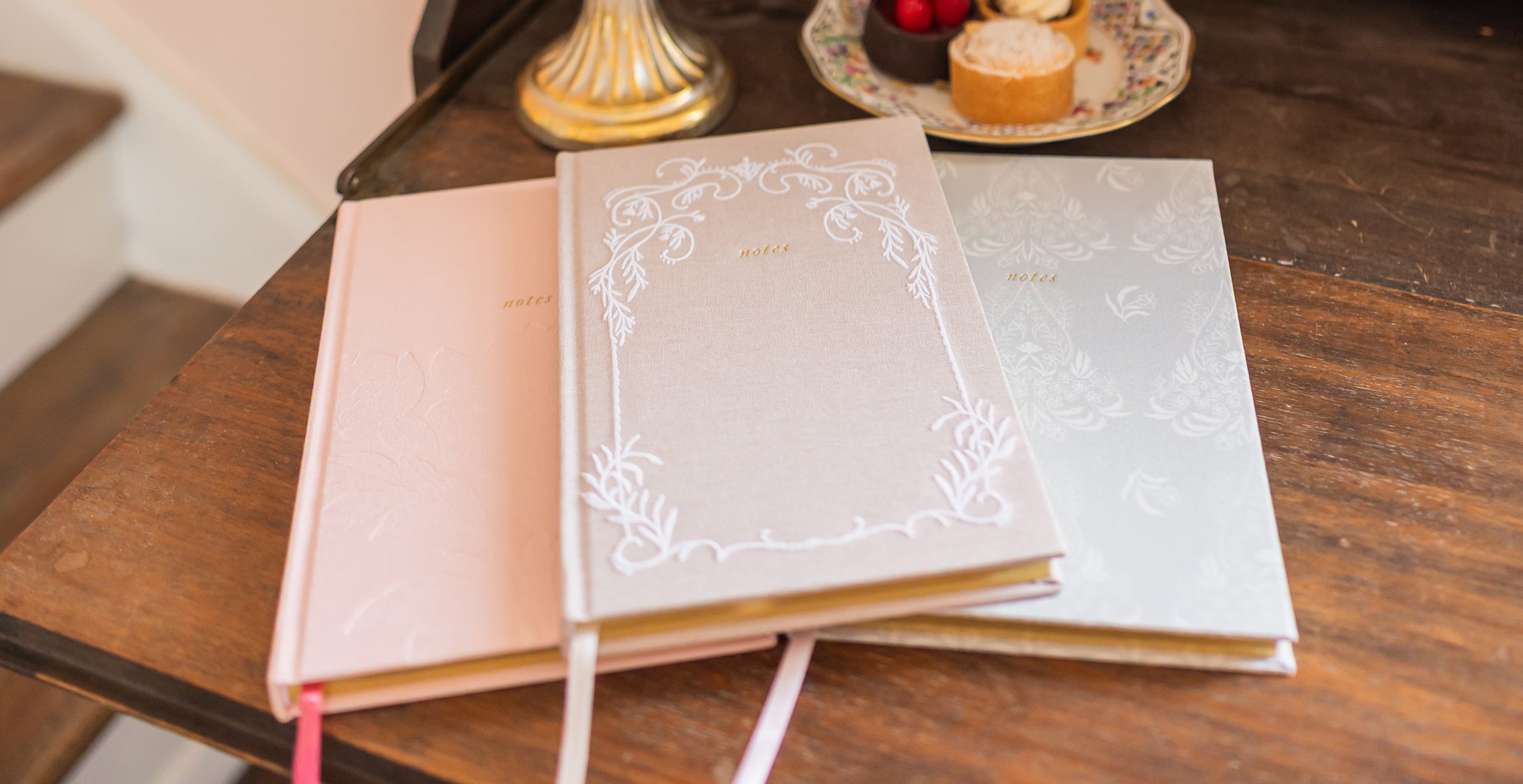Decorative setting with a vase of flowers, embroidered journals, and a plate of desserts on an antique desk.