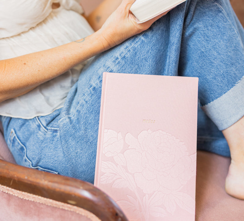 Person holding a pink notebook with floral debossing on a wooden chair.