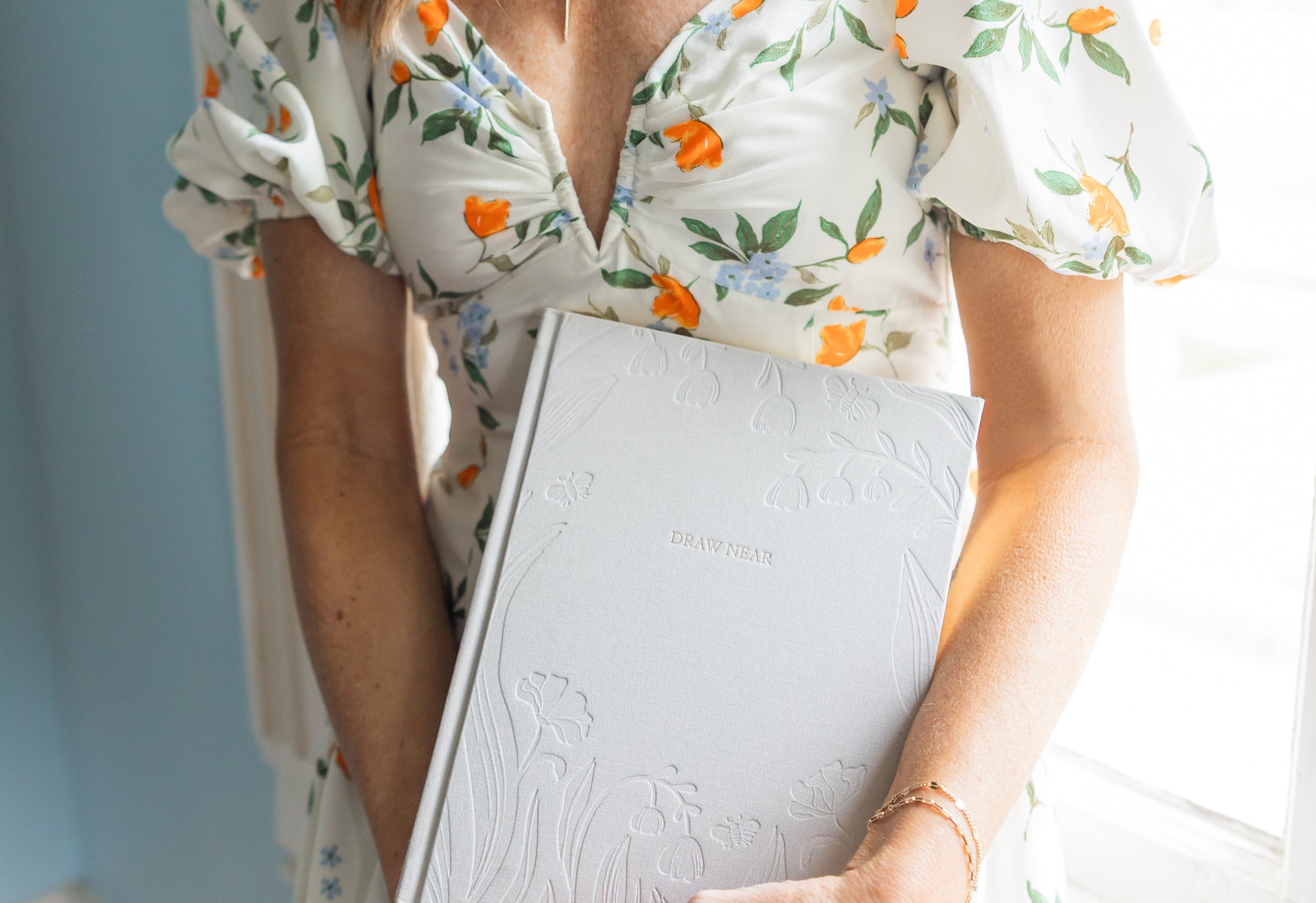Woman holding a Draw Near journal with a focus on the embossed details light blue wall in the background.