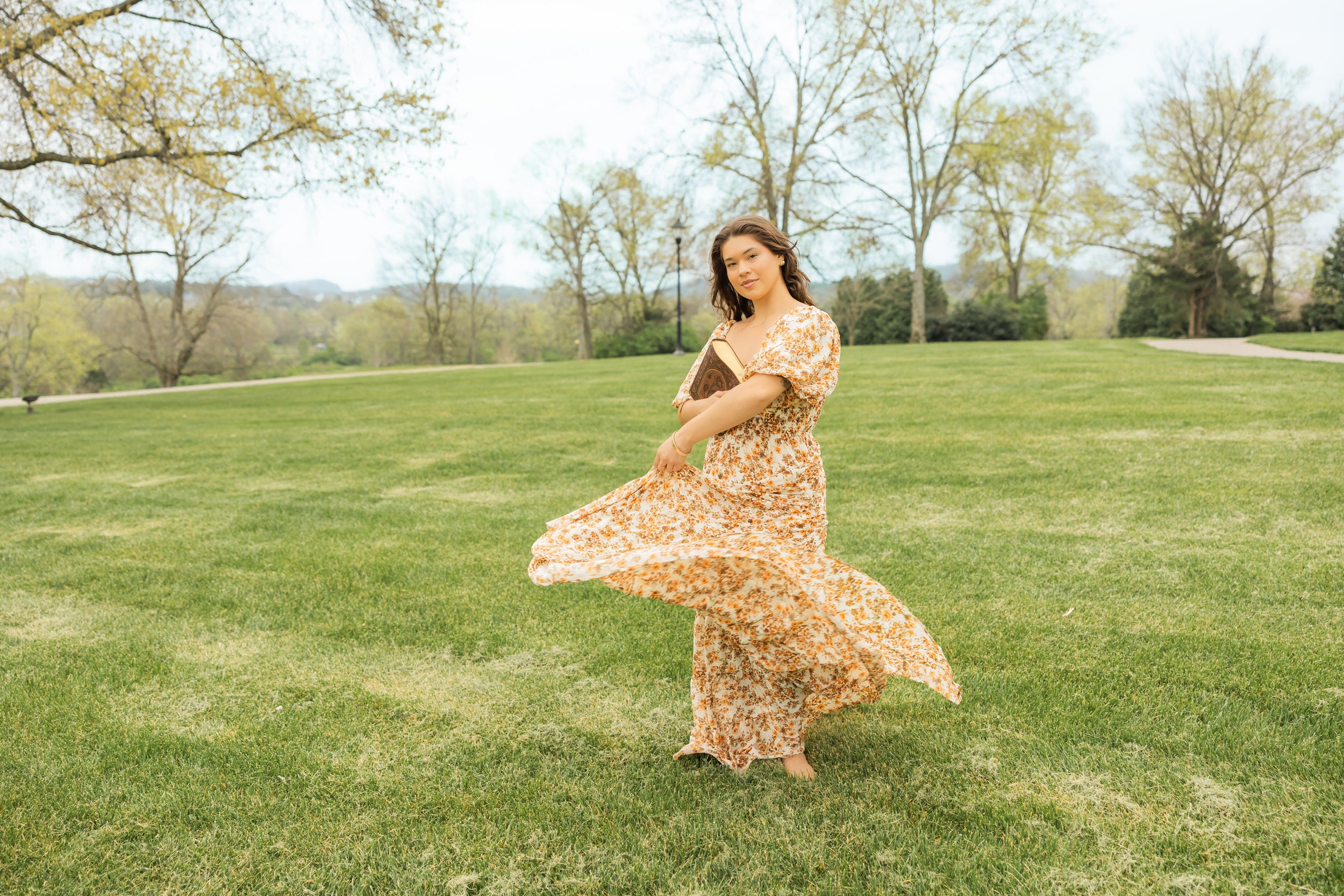 Woman in a floral dress standing on a grassy field with trees in the background, holding a Hosanna Revival bible.